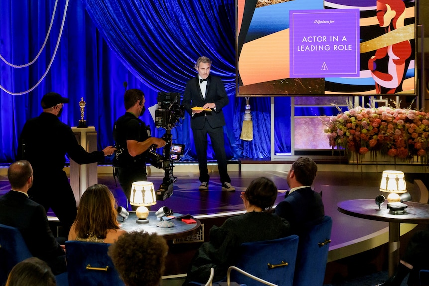 a man stands with note cards on a stage, two men are in front of the stage filming him