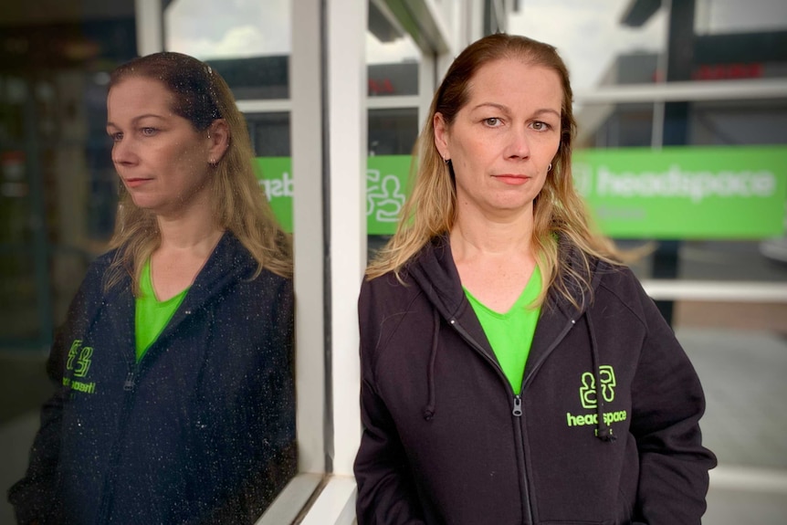 A woman with long blonde hair looks forward, a green Headspace sign is behind her, her refection can be seen in a glass window.