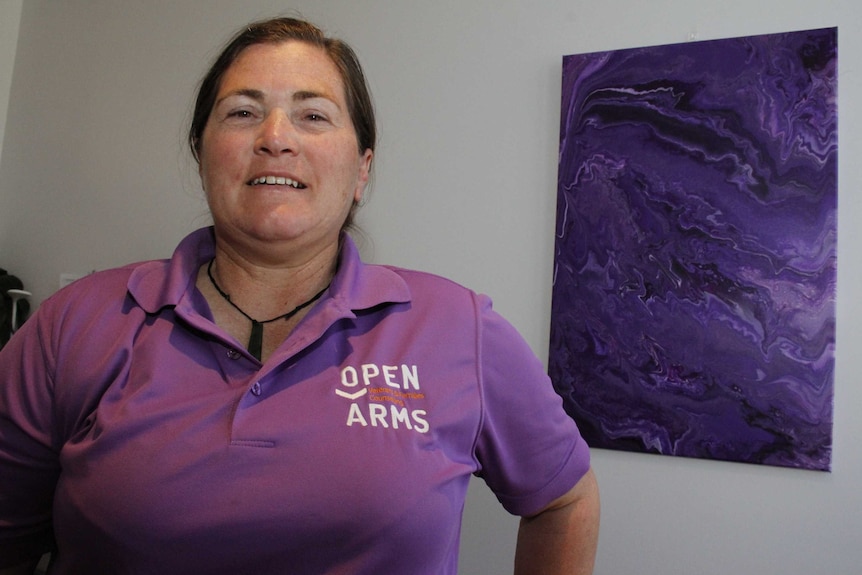 Woman in shirt with Open Arms logo standing in front of purple picture on wall.