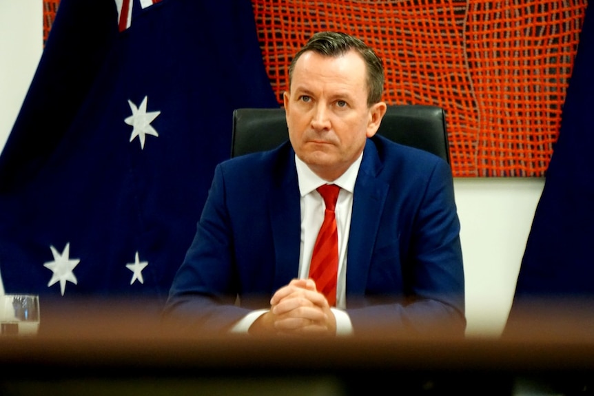 WA Premier Mark McGowan pictured seated at the head of a boardroom table with a flag behind him.