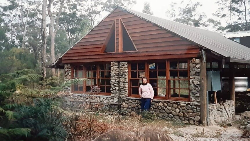 A person stands in front of a stone and timber house surrounded by bushland.