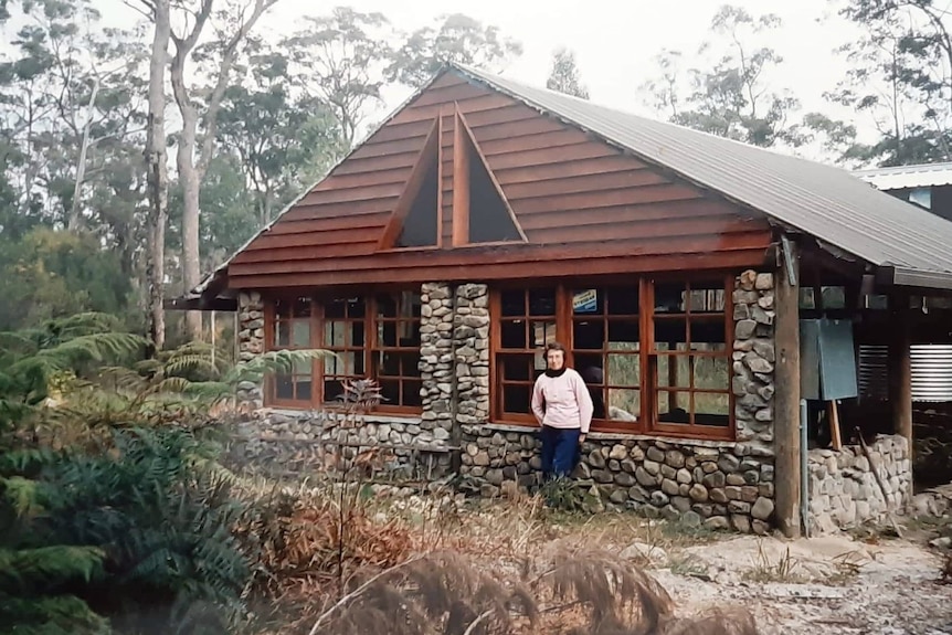 A person stands in front of a stone and timber house surrounded by bushland.