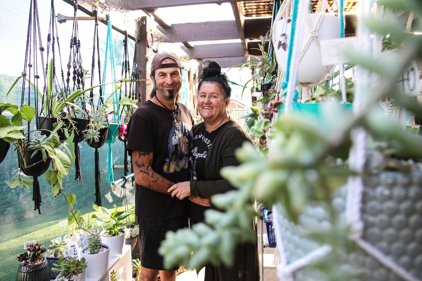 A couple wearing black t shirt stand amongst hanging pot holders with plants.