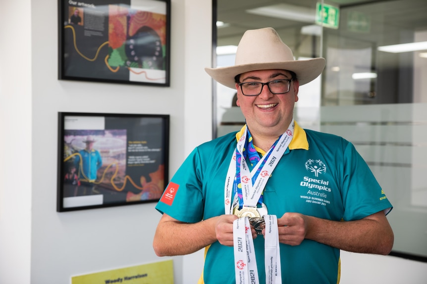 A young man wears the Australian Olympic polo shirt and a cowboy hat while holding medals and smiling