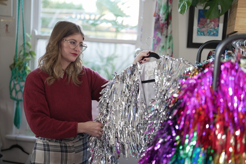 A young woman in a red jumper and large round glasses picking up a sparkly silver jacket from a clothing rail