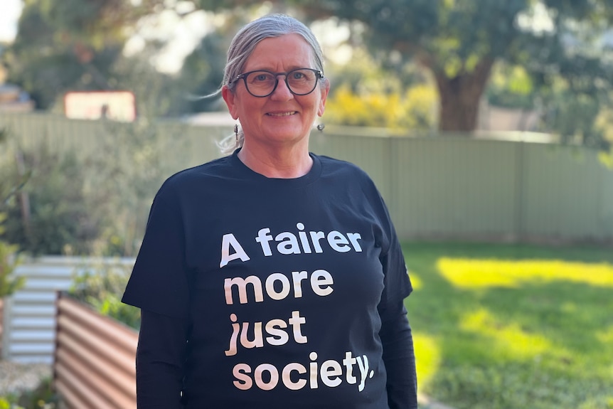 a woman with grey hair and glasses wears a t-shirt that reads a fairer and more just society