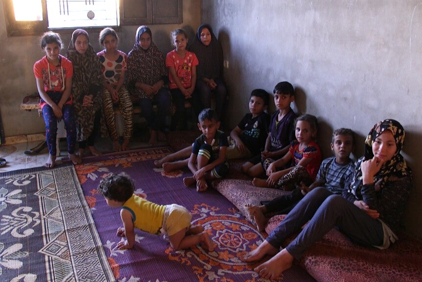 Six children sit on a bench and seven children sit on the floor of a small room