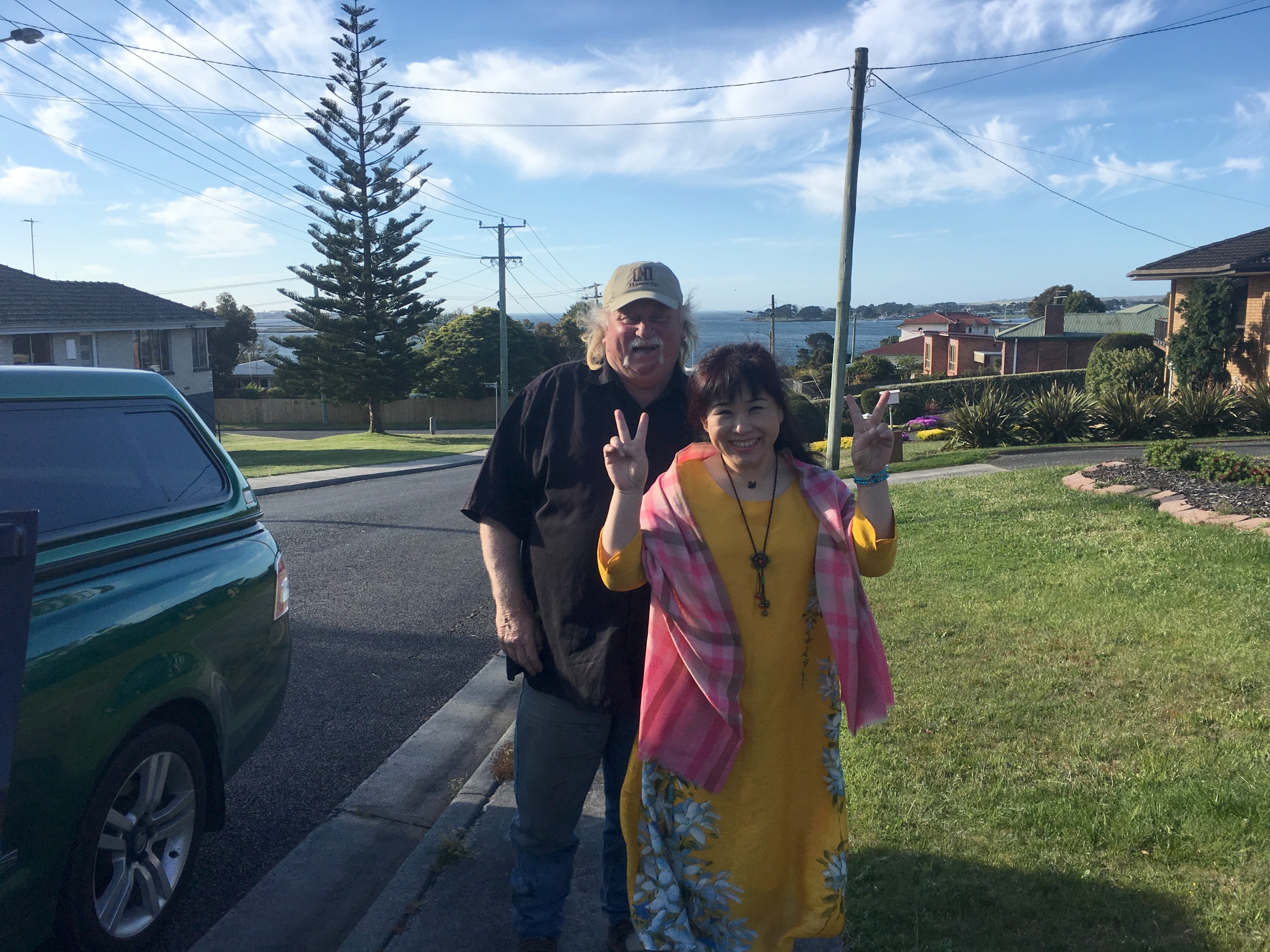 一位白发男子戴着棒球帽，面带微笑地站在一位面带微笑的女子身后，女子用双手比出和平手势。