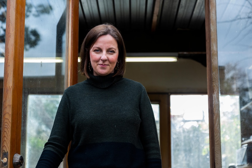 A woman with short brown hair and a dark jumper standing in a timber door frame.