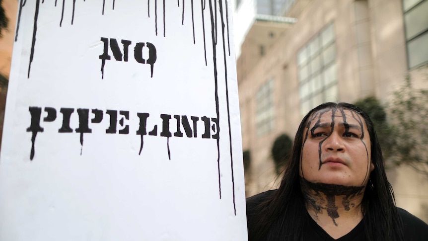 A Dakota Access Pipeline protester with black paint dripping down his face holds a sign saying "No Pipeline".