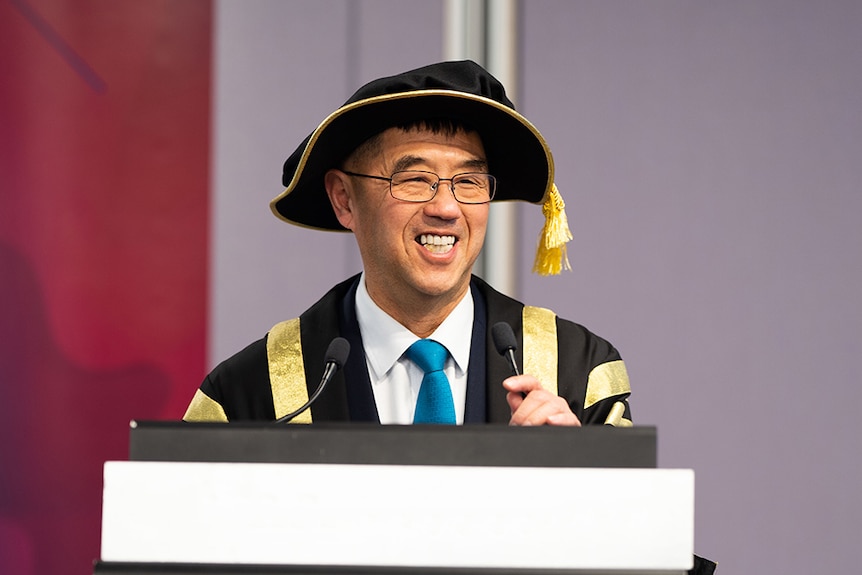 A man in academic dress smiles while speaking at a lectern.