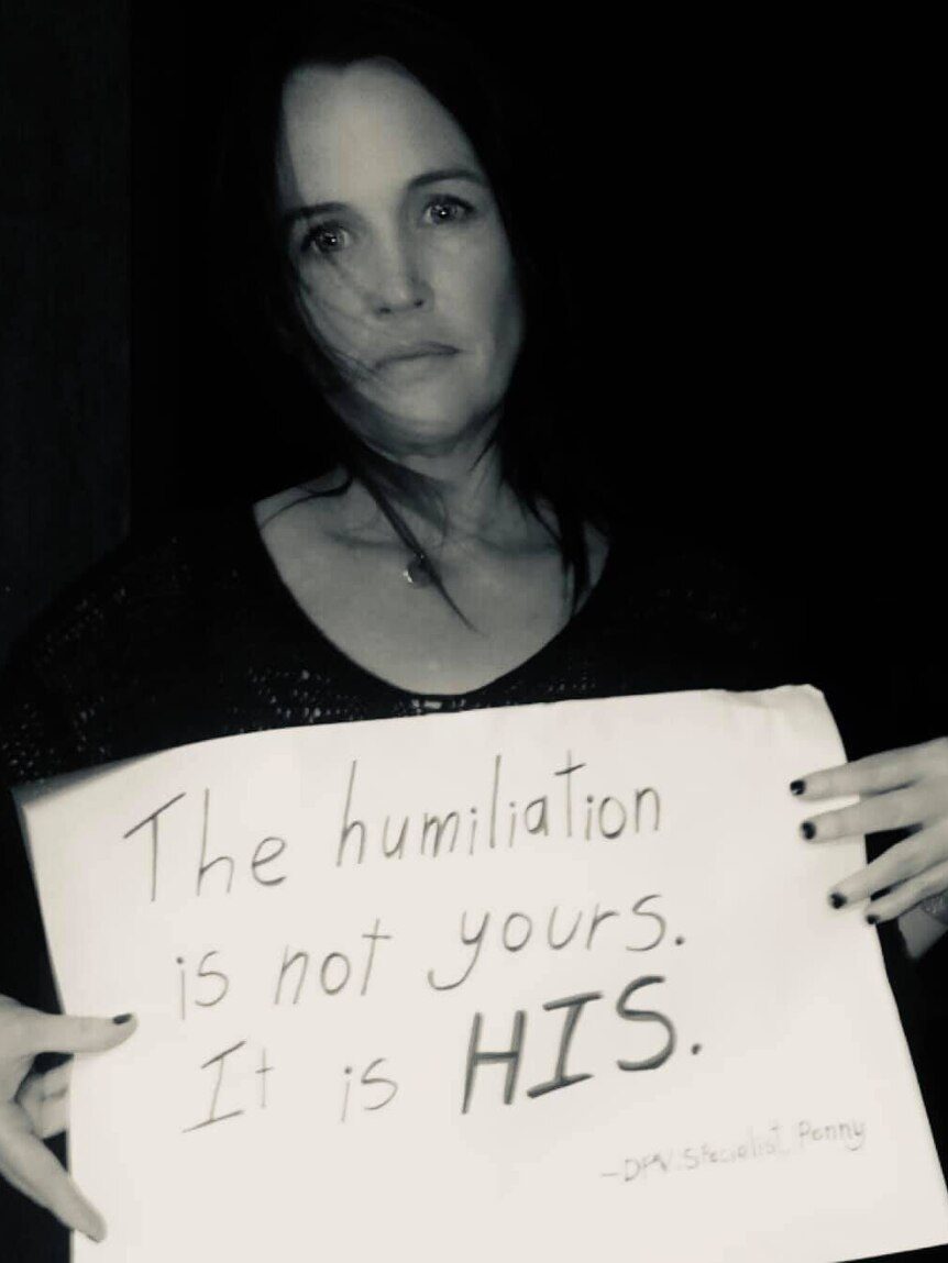 a black and white photo of a woman holding a placard saying "the humiliation is not yours, it is his"