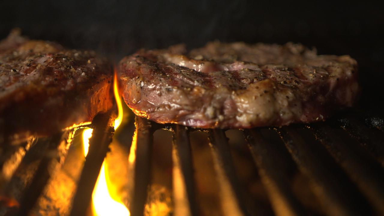 A close up of two pieces of steak on a barbecue grill. A small flame is rising through the grill.
