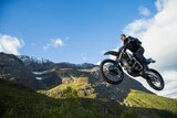 Tom Cruise rides a motorbike. He is in the air with mountains and blue sky in the background.