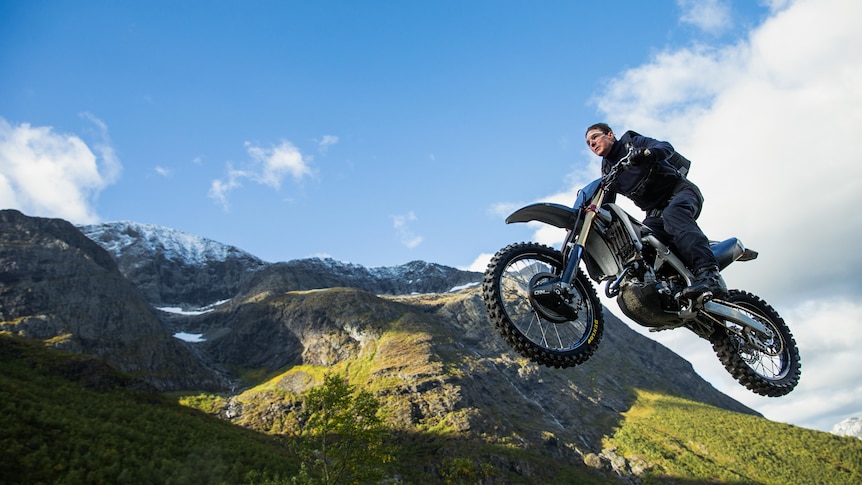 Tom Cruise rides a motorbike. He is in the air with mountains and blue sky in the background.