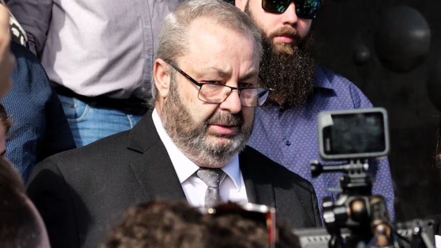 An older, bearded man with grey hair stands amid a crowd of journalist.