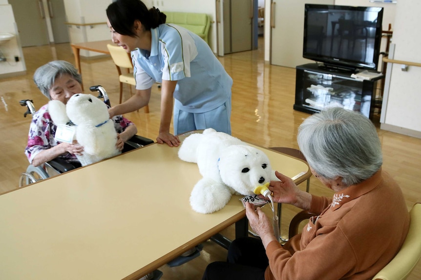 Two elderly women sit while playing with two robot seal plush toys.