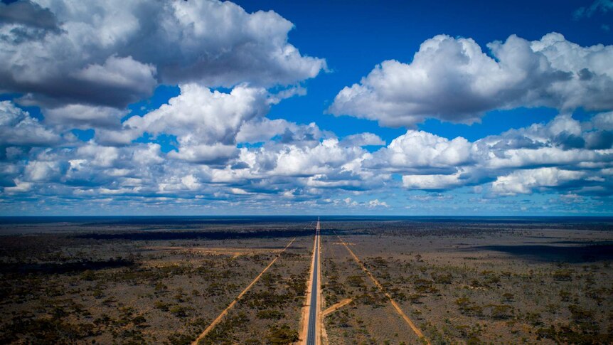 An aerial shot of of a blue sky with clouds, and a remote highway in cutting through outback scrubland. 