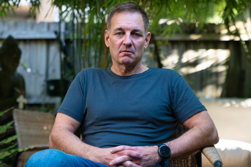 A man sits and looks at the camera in a garden. He has a sad expression. 