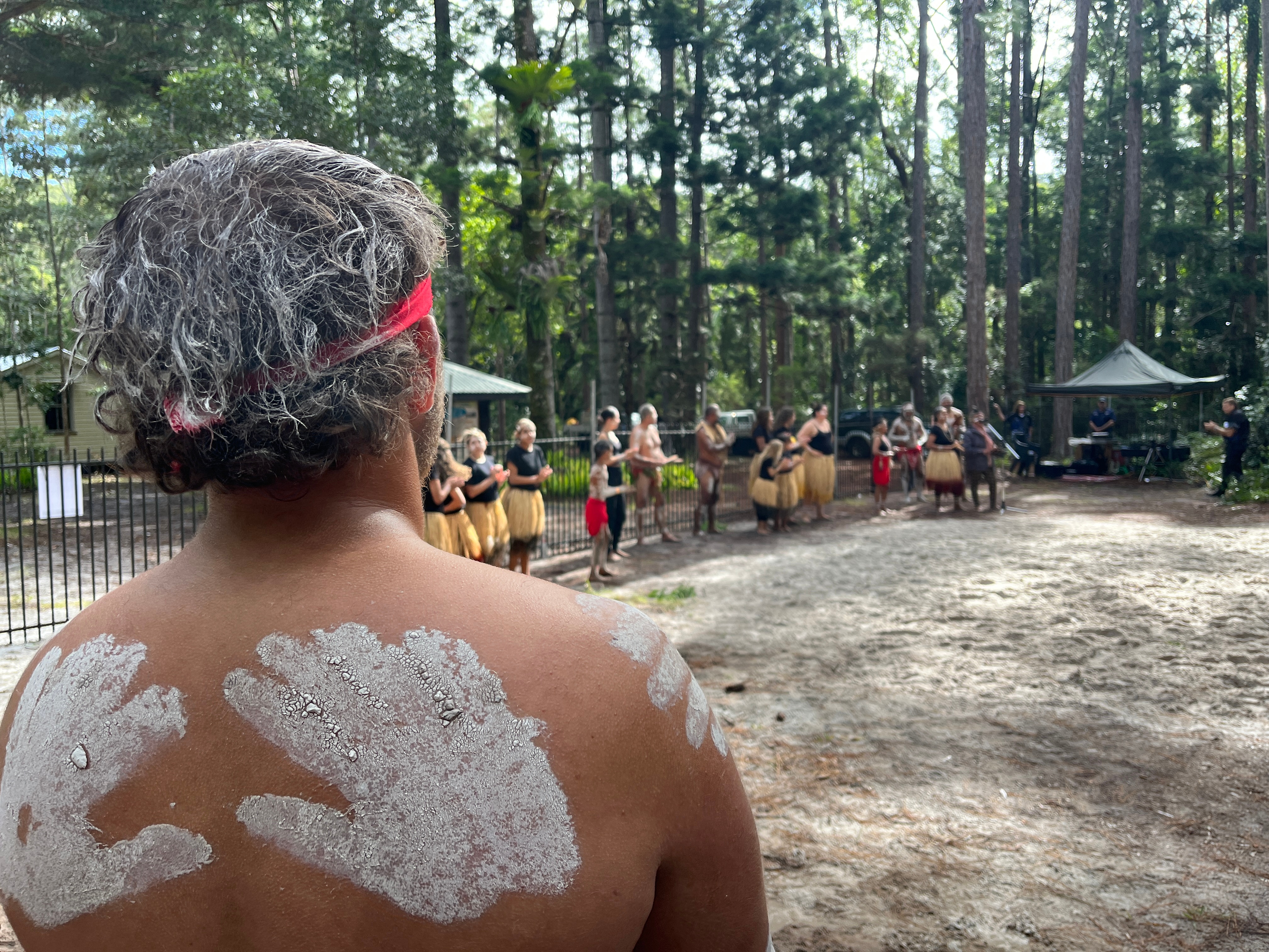The back of a man with clay finger prints looking out over a sandy area where people are standing in grass skirts