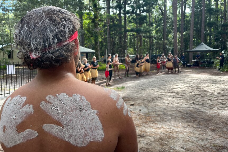 The back of a man with clay finger prints looking out over a sandy area where people are standing in grass skirts