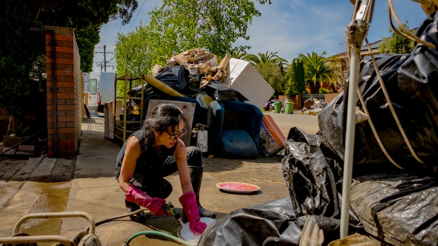 A woman cleans plates on the pavement as rubbish piles up around her