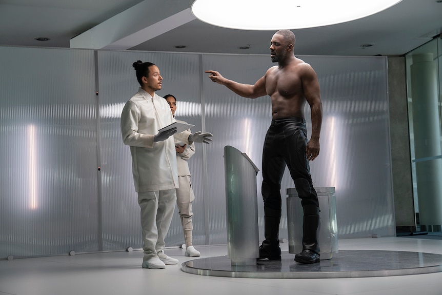 A topless muscular man stands and points at two people in lab coats in minimal and futuristic room.
