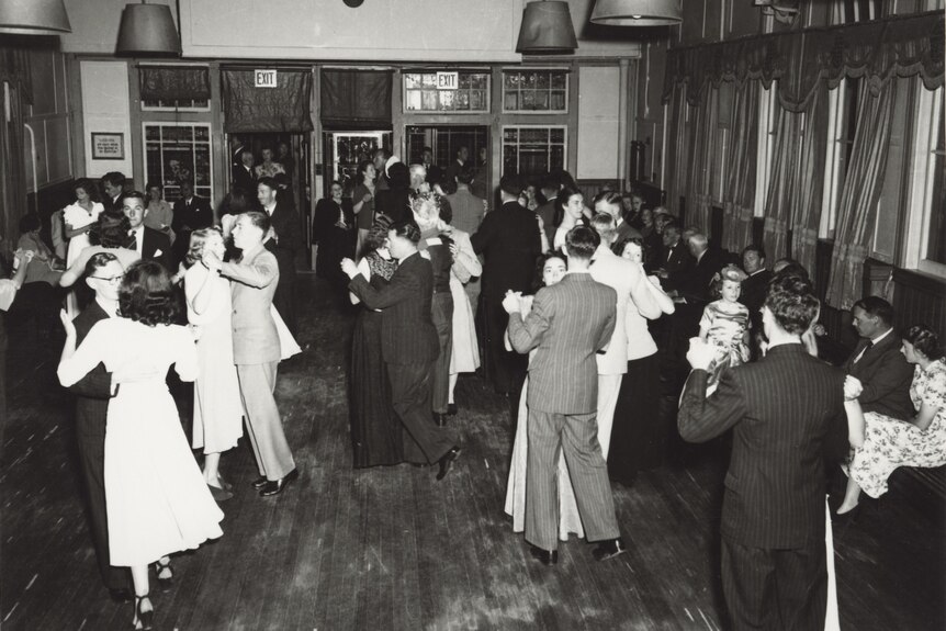 a black and white picture of a busy ballroom with partners dancing in suits and dresses