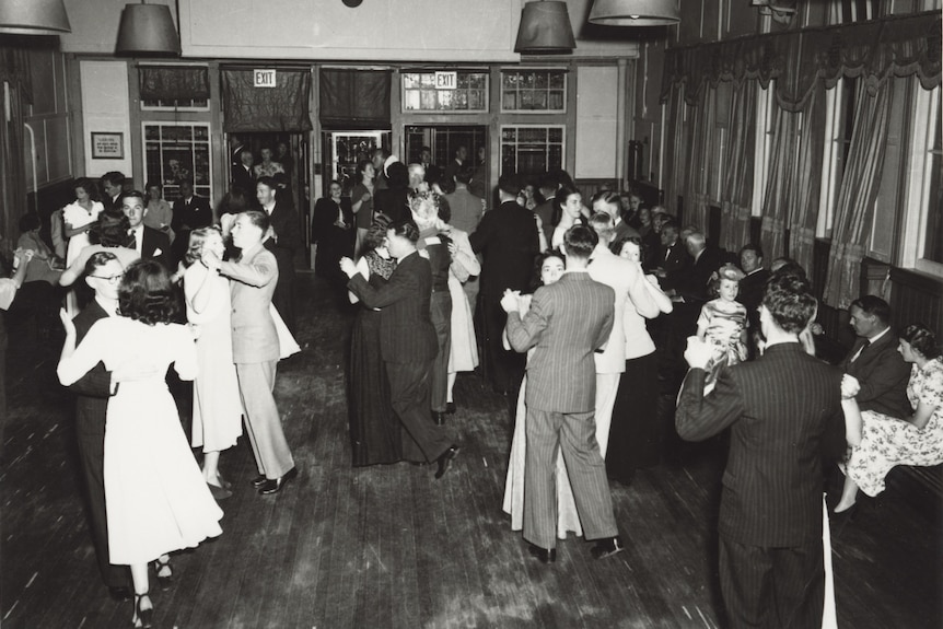 A black and white picture of a busy ballroom with partners dancing in suits and dresses.