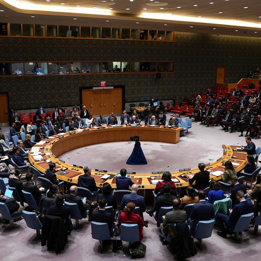 Diplomats sit around a large, circular table. More people sit to the side in tiered rows of chairs.