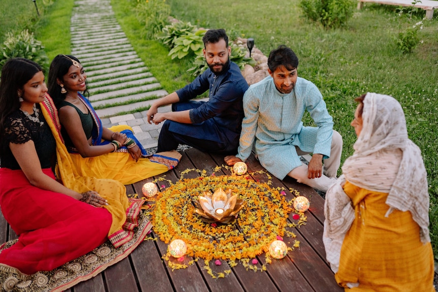People sitting on the ground, around a rangoli design made up of powder, flowers, rice and clay lamps.