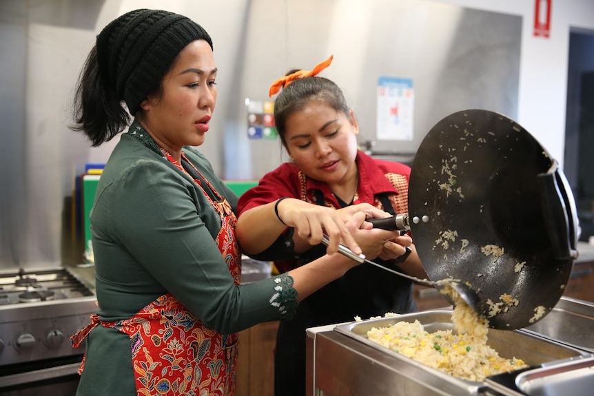 Two women dressed in traditional Thai outfits pour rice from a wok into a bain marie.