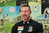Nathan Buckley sits behind a desk and a microphone, answering questions from journalist.