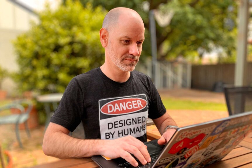 a man working on his laptop on a table, his shirt reads "danger, designed by humans"