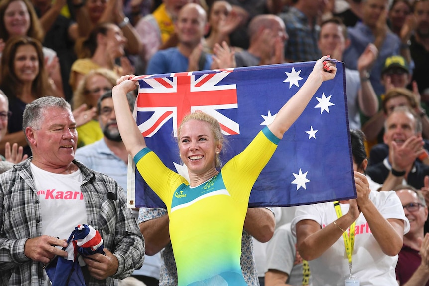 A young blonde athlete in a green and gold bodysuit holds the Australian flag aloft in front of an enraptured crowd.