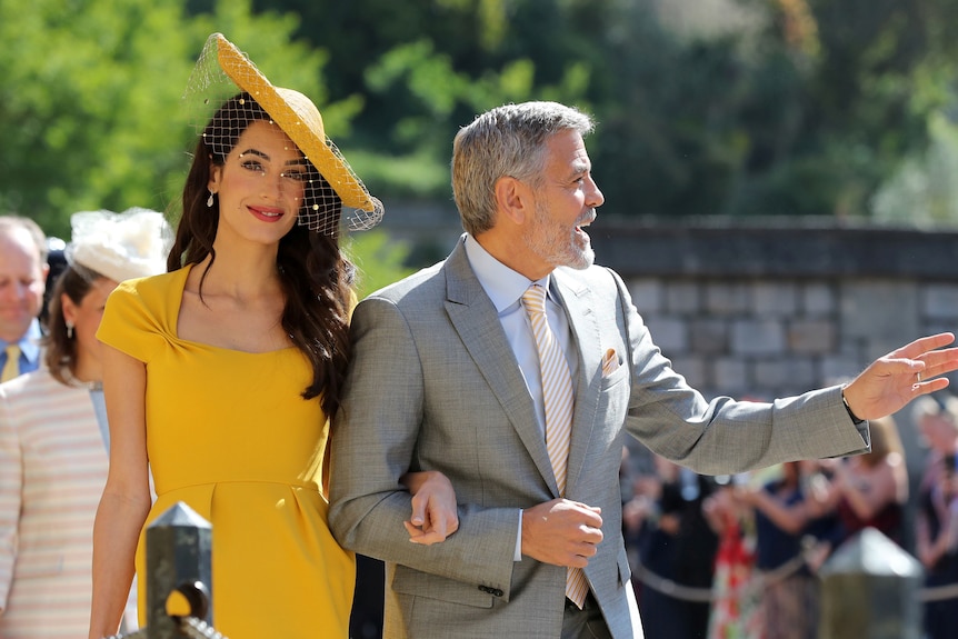 A woman in a yellow dress and matching hat smiles while a silver-haired man talks to someone out of frame