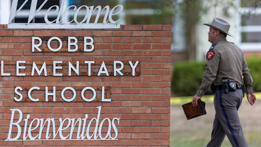 A police officer walks past a sign for Robb elementary school with Welcome above and Bienvenidos below