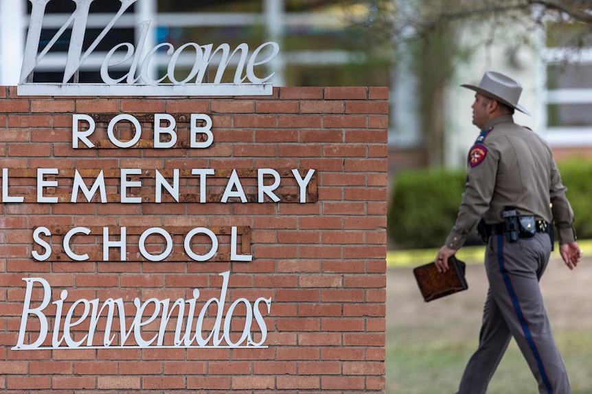 A police officer walks past a sign for Robb elementary school with Welcome above and Bienvenidos below