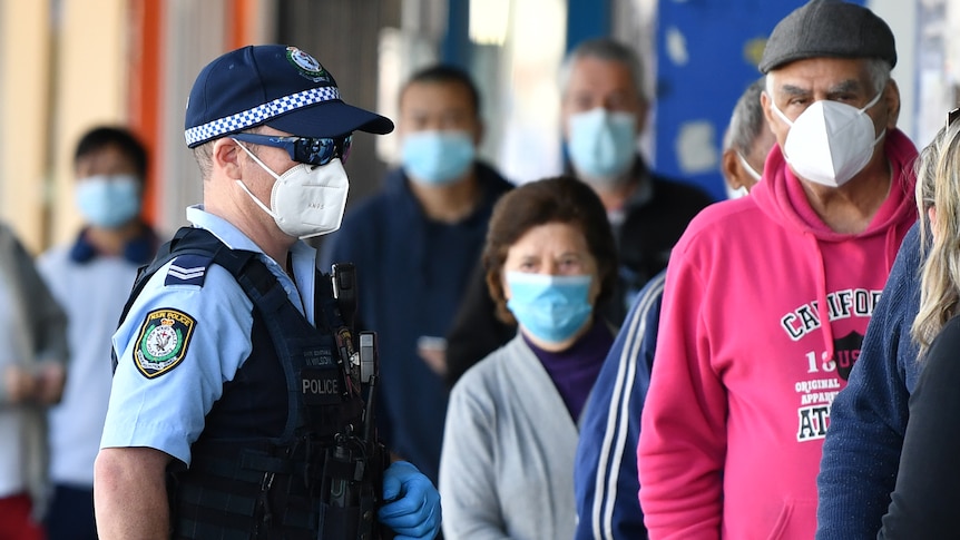Members of the public wearing face masks queue outside a bank as a police officer watches. 