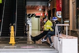 A man in high vis and a woman in a bike helmet sit looking at their phones and wearing face masks in a deserted shopping centre.