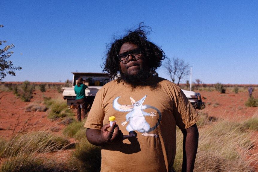 An Aboriginal man in a bilby shirt holds a sample vial on a red dusty spinifex plain in front of a ute