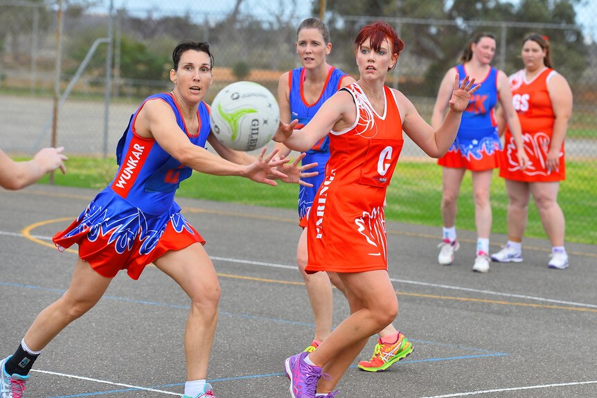 Two women, one wearing a blue netball uniform and another a red uniform compete to catch a ball. 