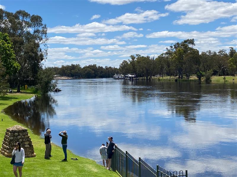 A small gathering of people stand near a swollen river on a sunny day