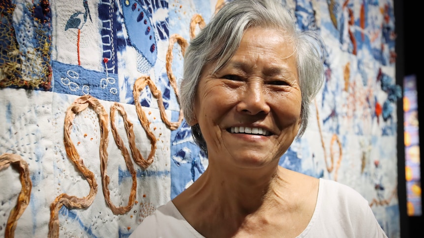 A portrait of a lady standing in front of a quilt.