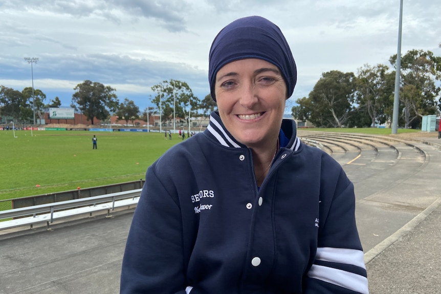 A woman in a headpiece and dark jacket sitting on the sidelines of a footy field