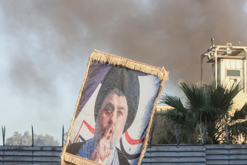 A protester holds up a portrait of Iraqi politician Moqtada al-Sadr as smoke rises in the background. 