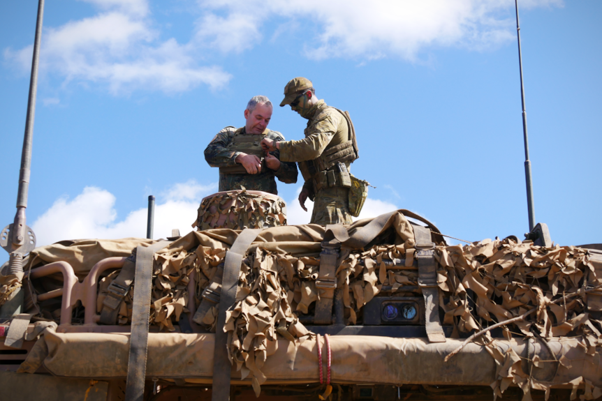 A soldier helps another man adjust his body armour while on the roof of an army vehicle