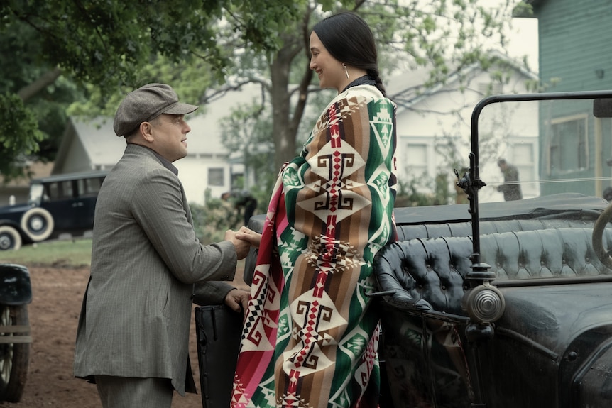 A film still showing a white man in a suit and cloth hat kneeling before a Native American woman wearing a traditional shawl