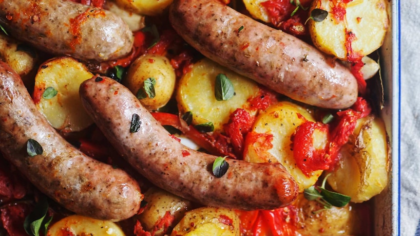 A tray with baked potato slices, crushed tinned tomato, capsicum, pork sausages and oregano, a tray bake dinner recipe.