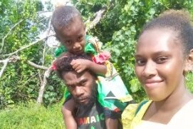 A Ni-Vanuatu mother and father walk together outside with child on his shoulders